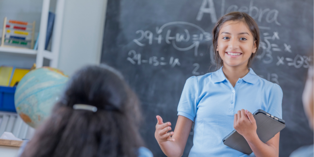 A girl in school uniform talks to class . she has a smile and a book